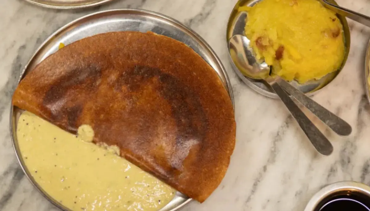 Masala dosa on a metal plate with coconut chutney on the side and potato curry nearby on a marble table