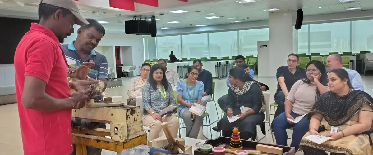 Instructor demonstrates pottery wheel technique to a diverse group of adults seated in a bright room at a workshop conference center, watching closely.