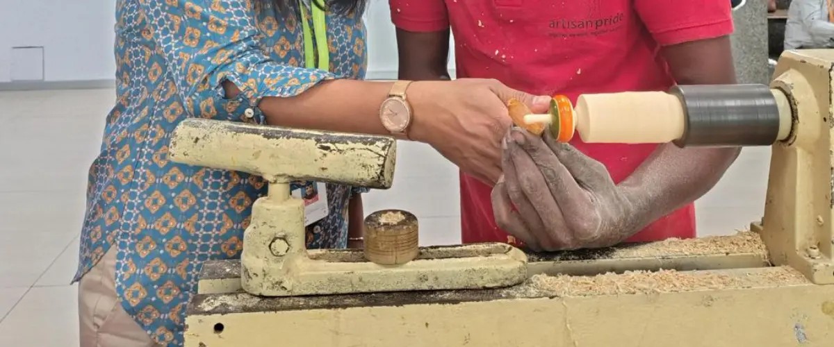 Two people work together at a wood lathe, shaping a wooden spindle as shavings fly nearby.