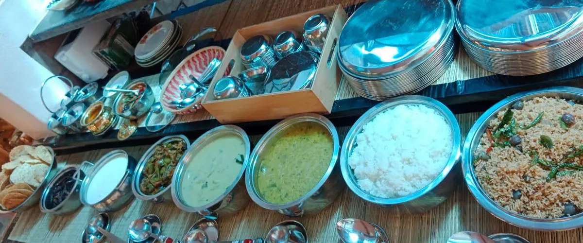 Buffet line with stainless steel chafing dishes containing rice, curries, and vegetables; plates and utensils nearby.