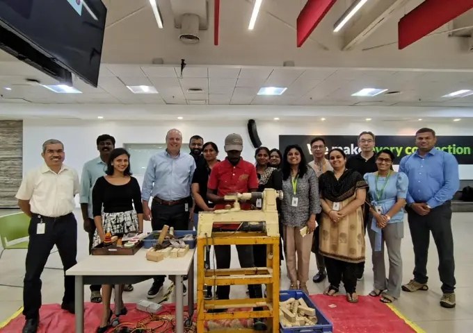 Group of colleagues posing around a wooden loom in a bright workshop classroom.