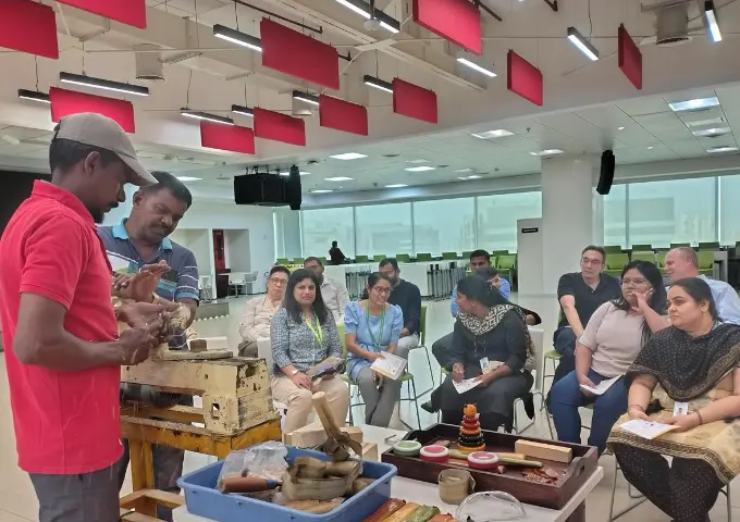 Instructor in a red shirt demonstrates wood carving on a lathe to a diverse group of coworkers in a modern office training room.