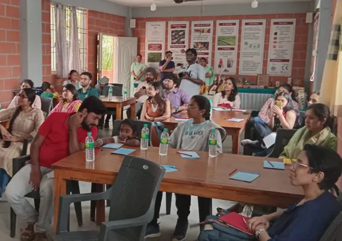 Group of adults and children in a classroom workshop, seated at tables with notebooks and water bottles, posters on the back wall.