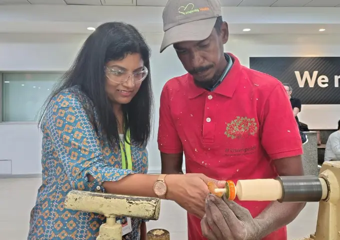 Two people wearing safety glasses watch and guide a turning wooden piece on a wood lathe during a workshop session.
