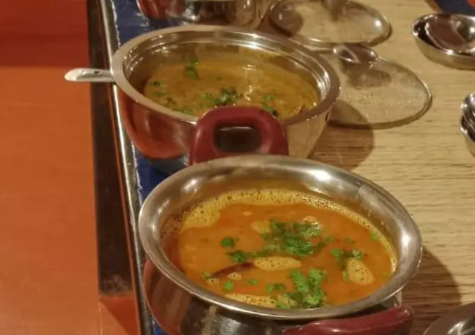 Two stainless steel bowls of curry with cilantro garnish on a wooden table in a restaurant setting.