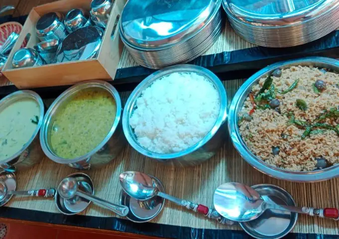 Buffet spread with metal bowls containing yellow curry, green lentil soup, steamed white rice, and a spiced grain dish; metal spoons laid out on the counter.