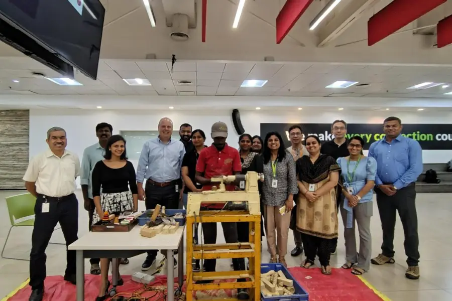 A diverse group of people standing in a workshop around a wooden lathe and wood pieces on a table inside a modern office space.