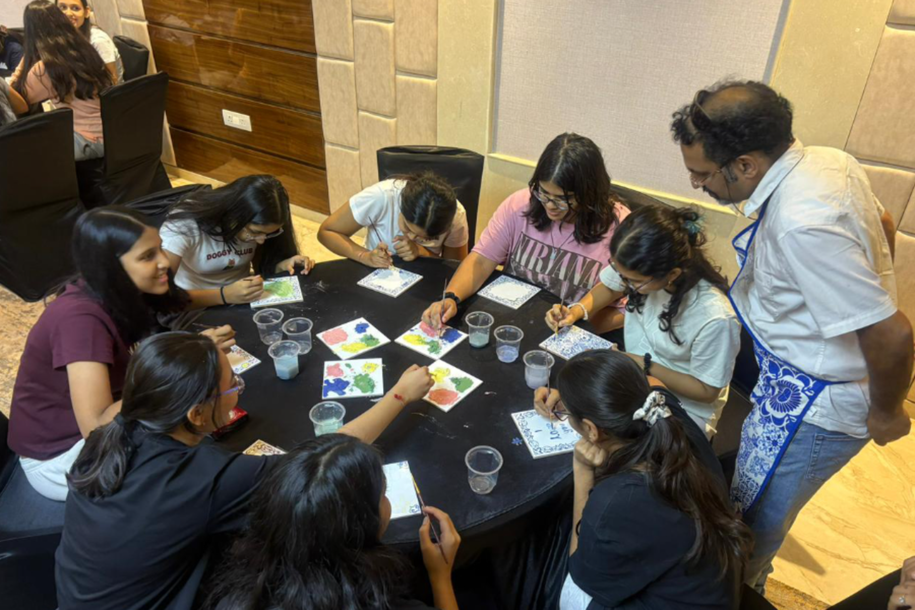 Group of students painting colorful tiles around a black table, instructor standing beside them in a studio.