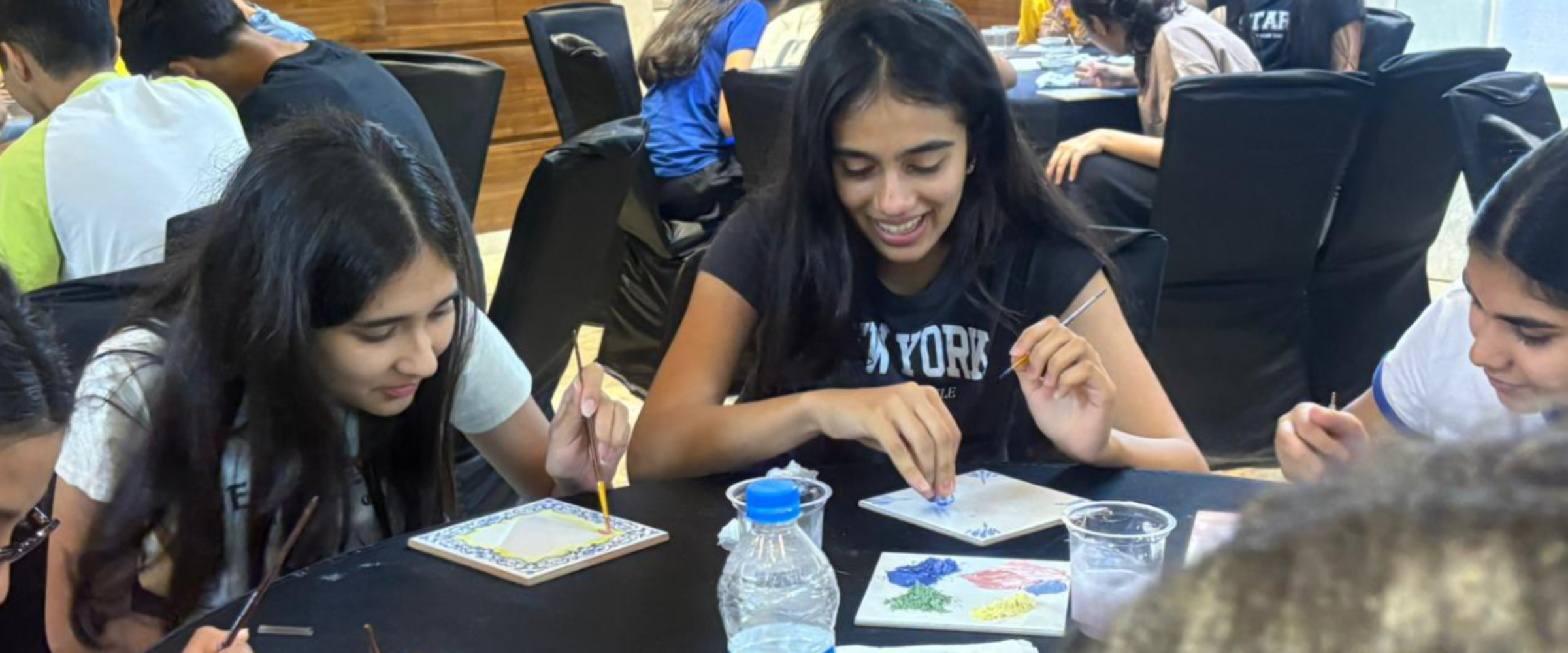 Teenagers painting small tiles at a long table during a group art activity in a bright room