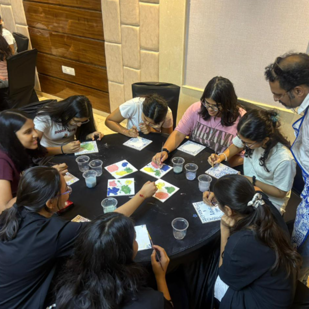 Group of people around a round table painting colorful tiles in a craft workshop, with cups of water nearby.