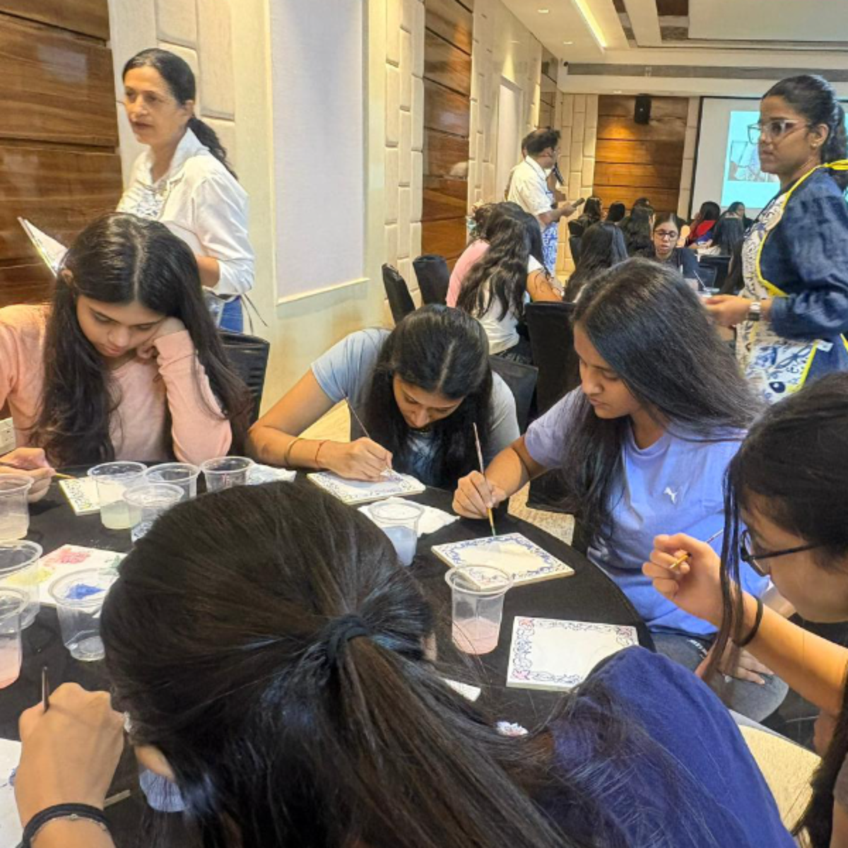Group of young women sitting around a round table painting small tiles with brushes in a bright conference room.