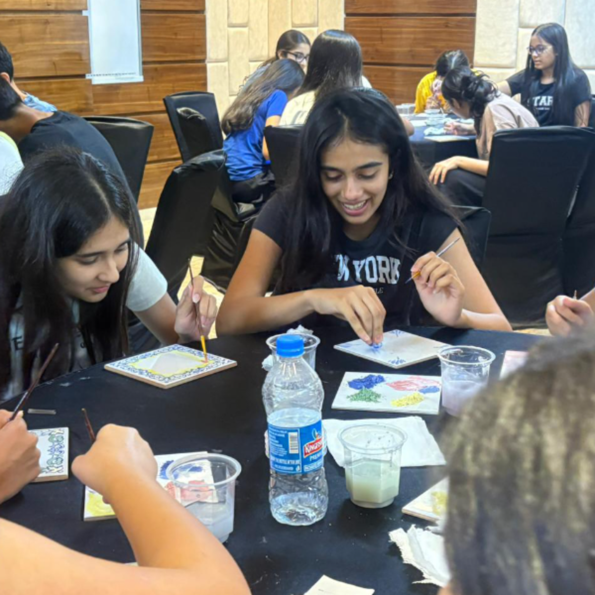 A group of students sits around a black table, painting colorful tiles with brushes and small cups of paint.