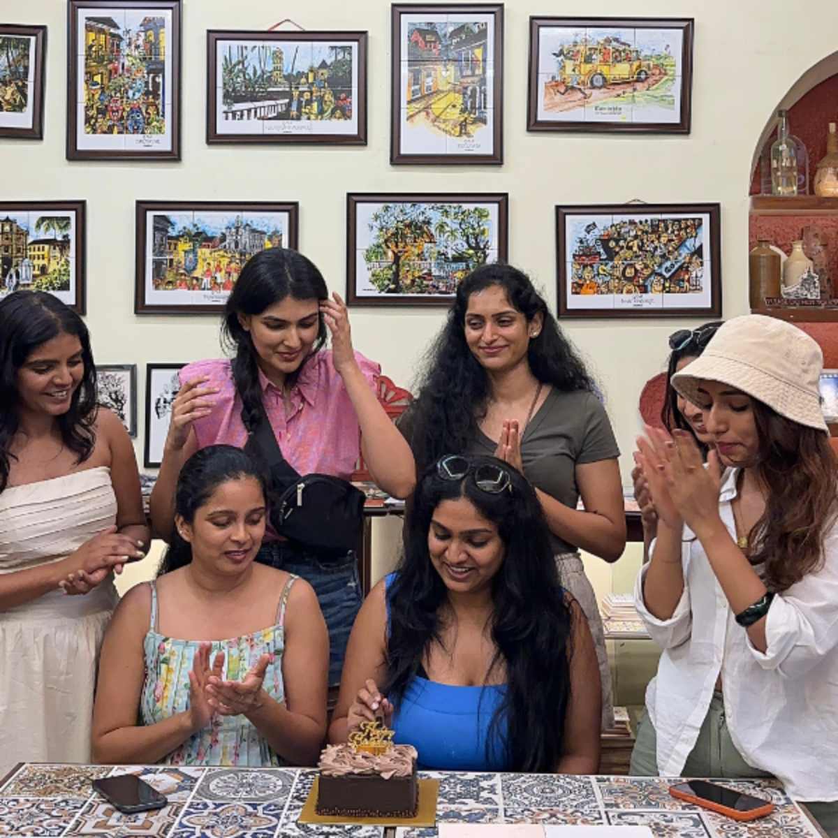 Group of six women gathered around a table, one woman about to cut a chocolate cake with decorative topping, others clapping and smiling in celebration in a bright room with framed art on the wall behind them.
