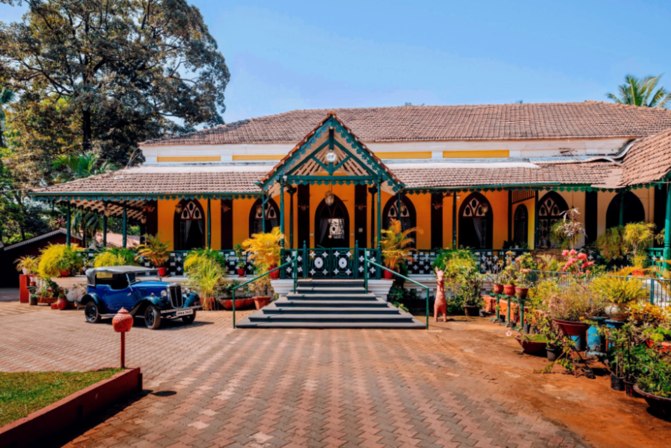 Yellow colonial building with green trim, arched windows, and steps leading to the entrance amid potted plants and a vintage blue car nearby.