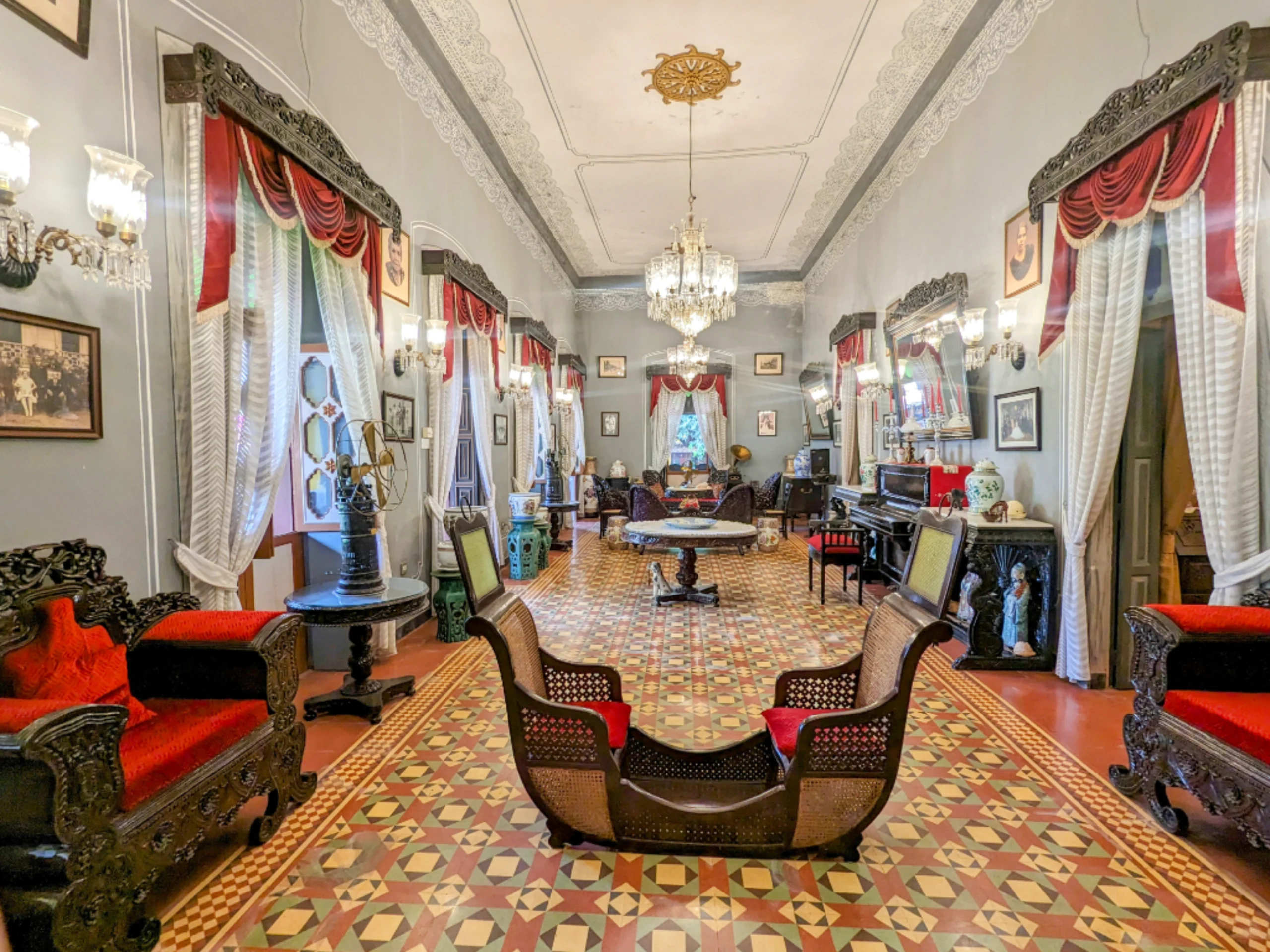 Luxurious Victorian-style living room with crystal chandeliers, dark carved furniture, and red and white draped curtains.