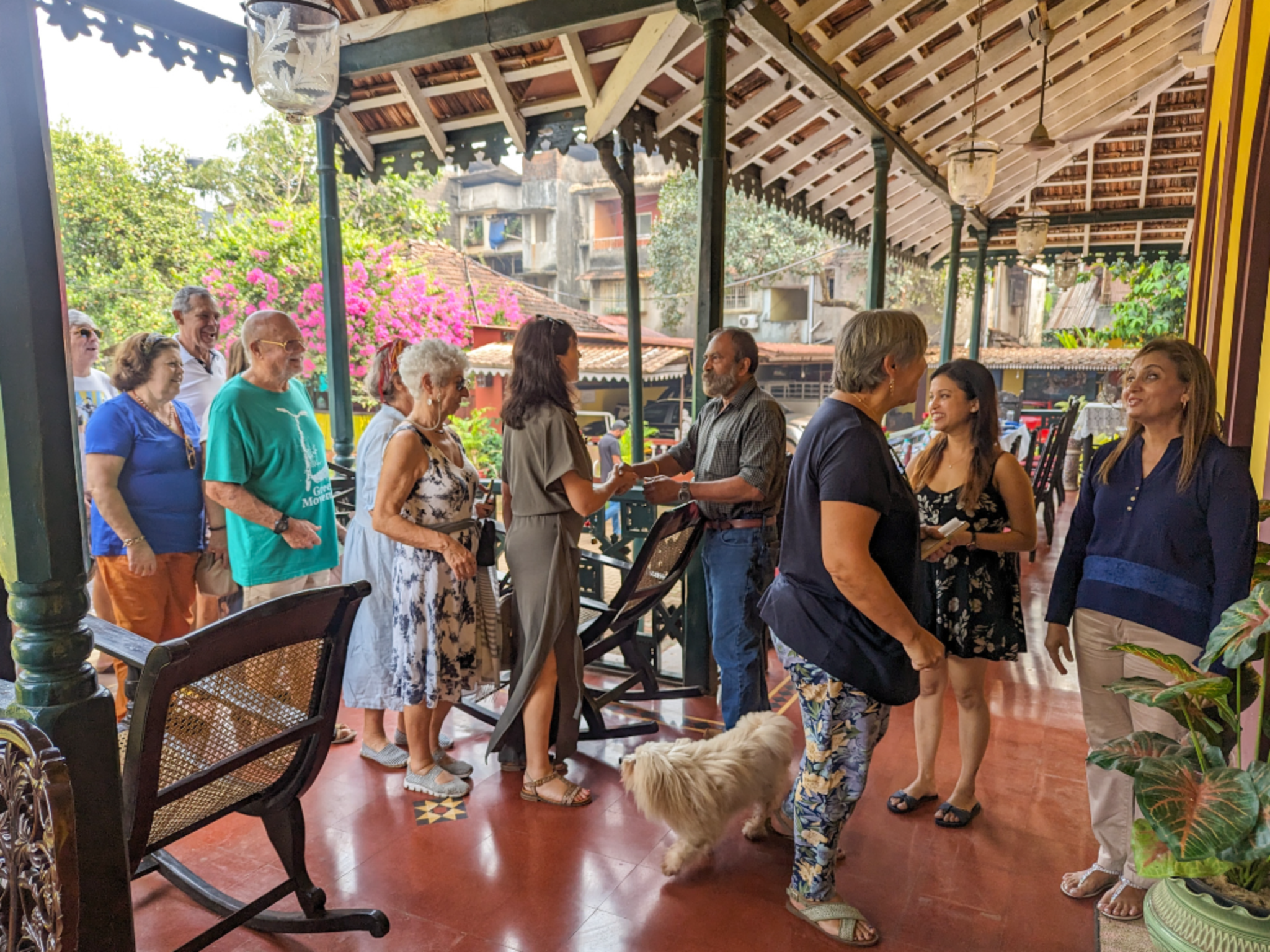 Group of people chatting and smiling on a covered outdoor porch, with a fluffy white dog at their feet and pink bougainvillea in the background.