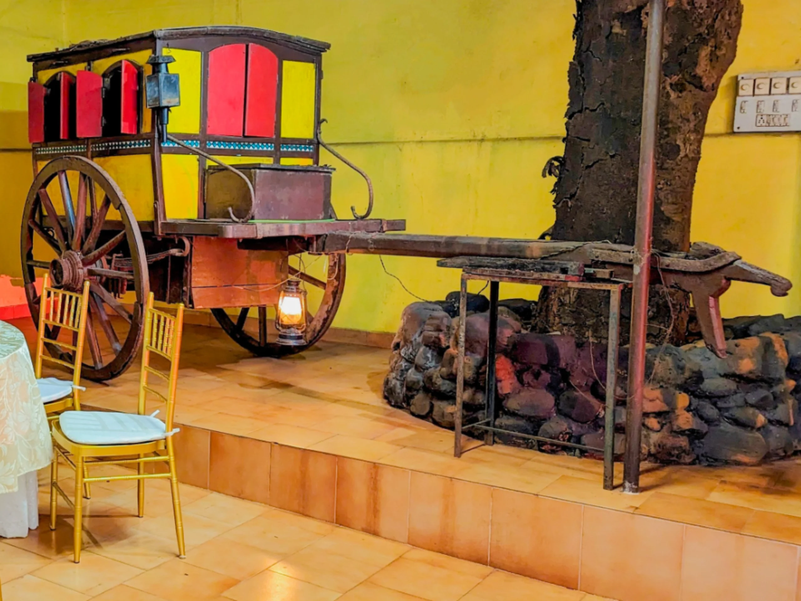 Old wooden cart with red compartments and a lantern indoors against a bright yellow wall, with yellow chairs nearby.