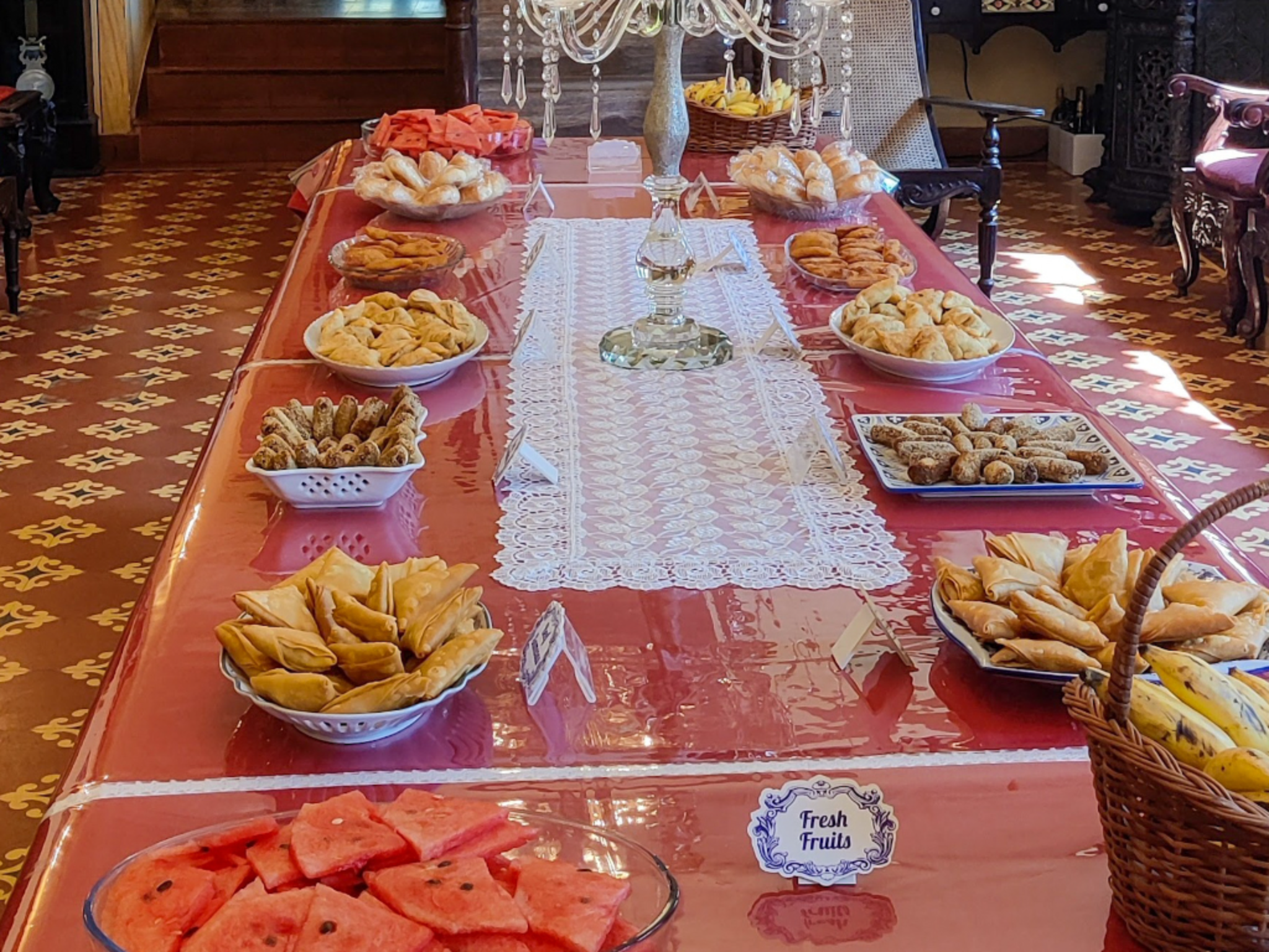 Buffet table with a pink tablecloth, bowls of pastries and sliced watermelon, labeled 'Fresh Fruits' in a formal dining room.