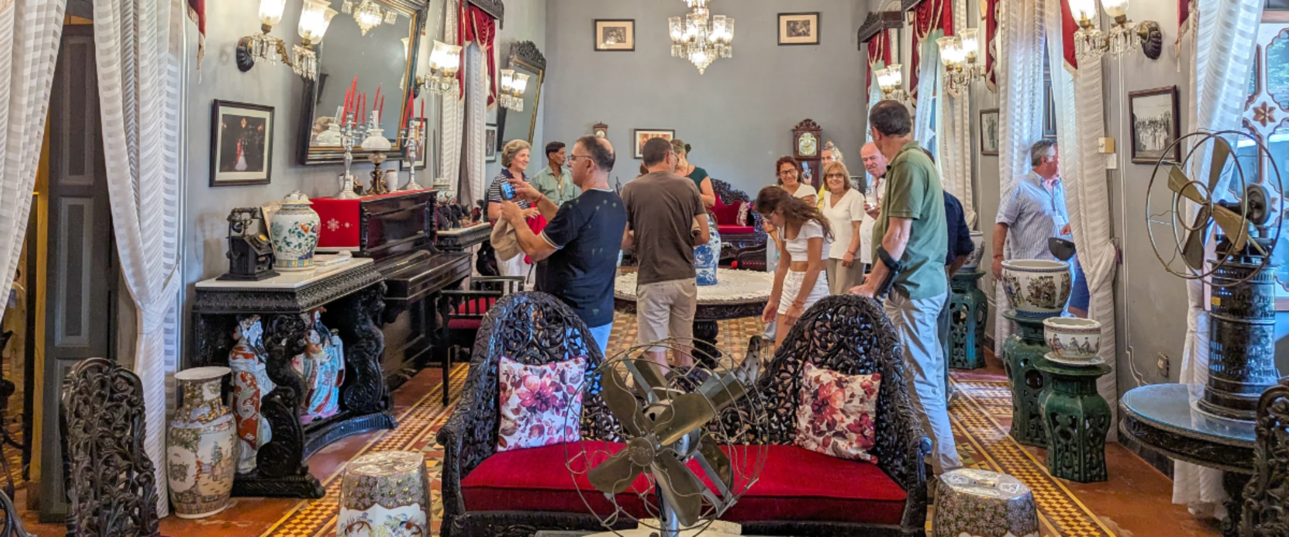 A group of people gathers around a black piano in a richly decorated antique showroom with chandeliers, vases, and ornate chairs.