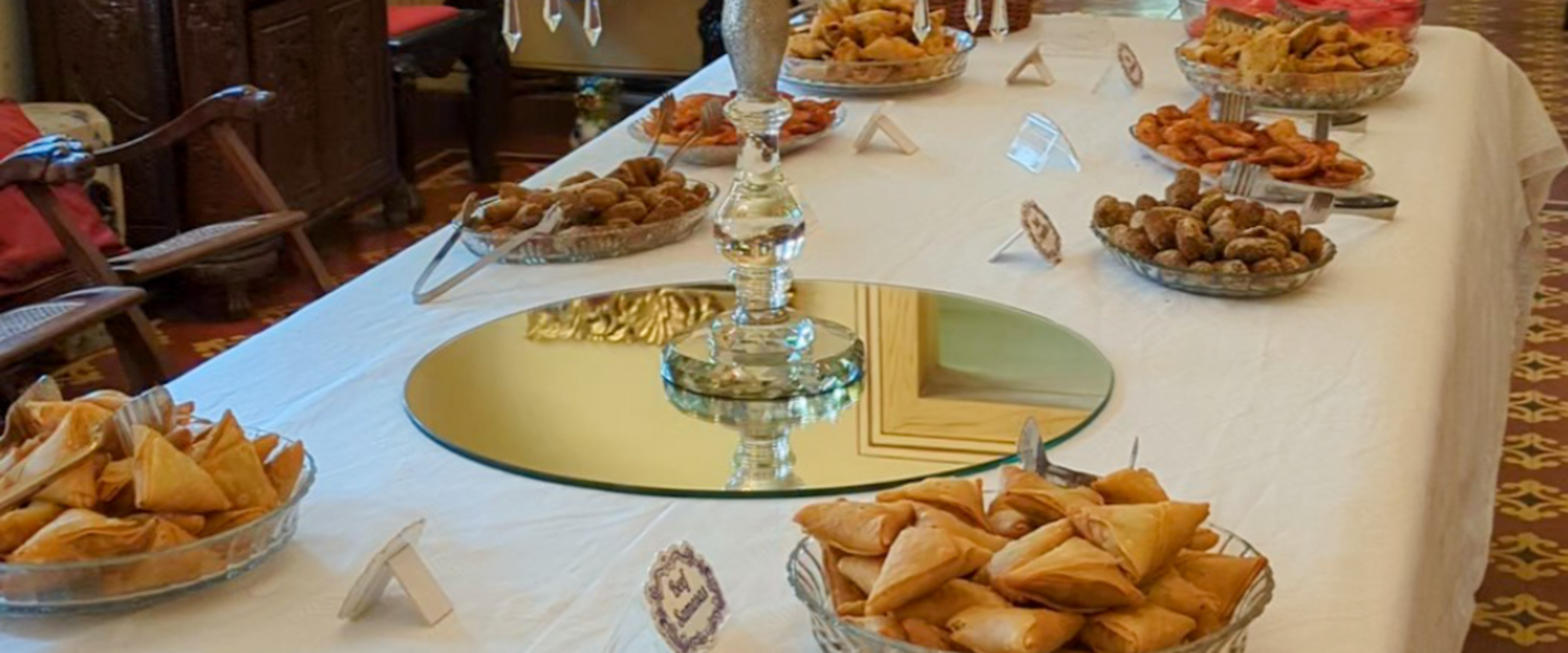 Assorted pastries and finger foods arranged on a long white-clothed banquet table at a formal event, with a mirrored centerpiece.