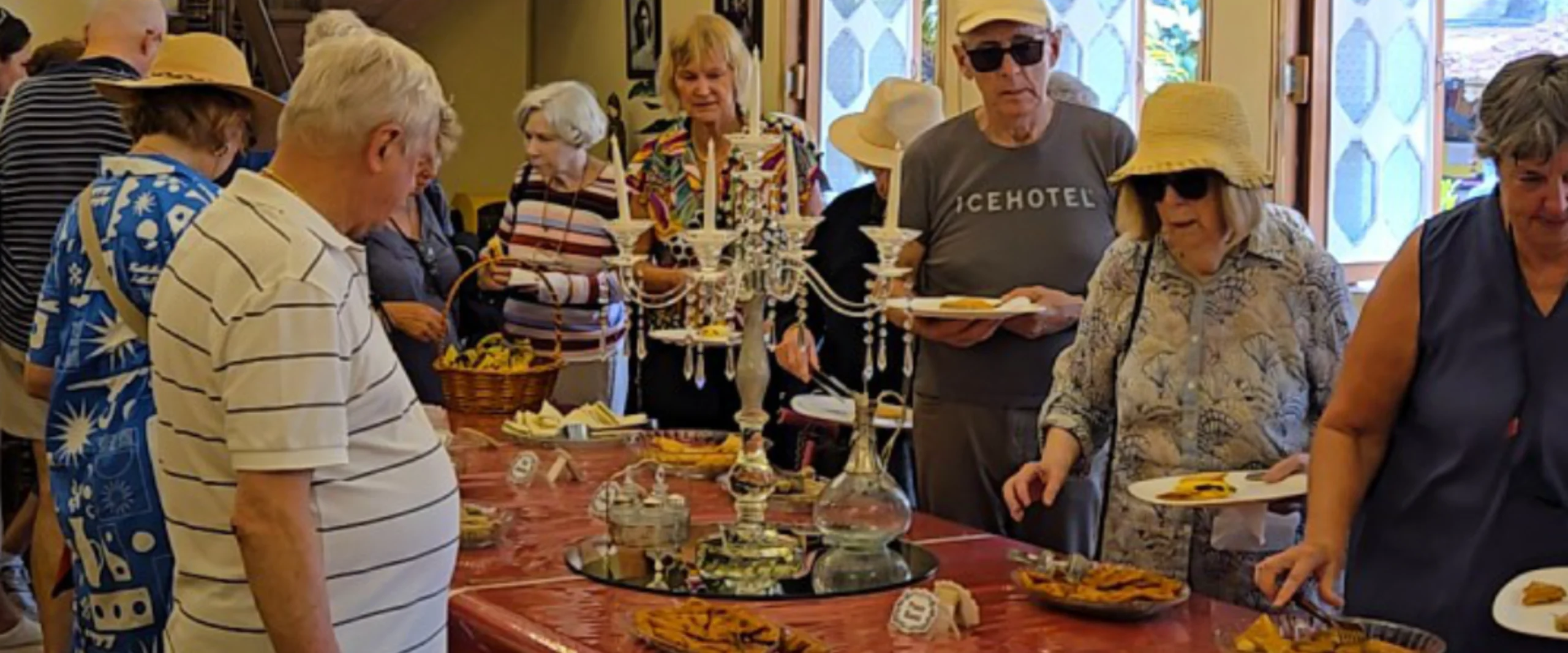Group of seniors gathered around a buffet-style table with pastries and a crystal centerpiece in a bright room.