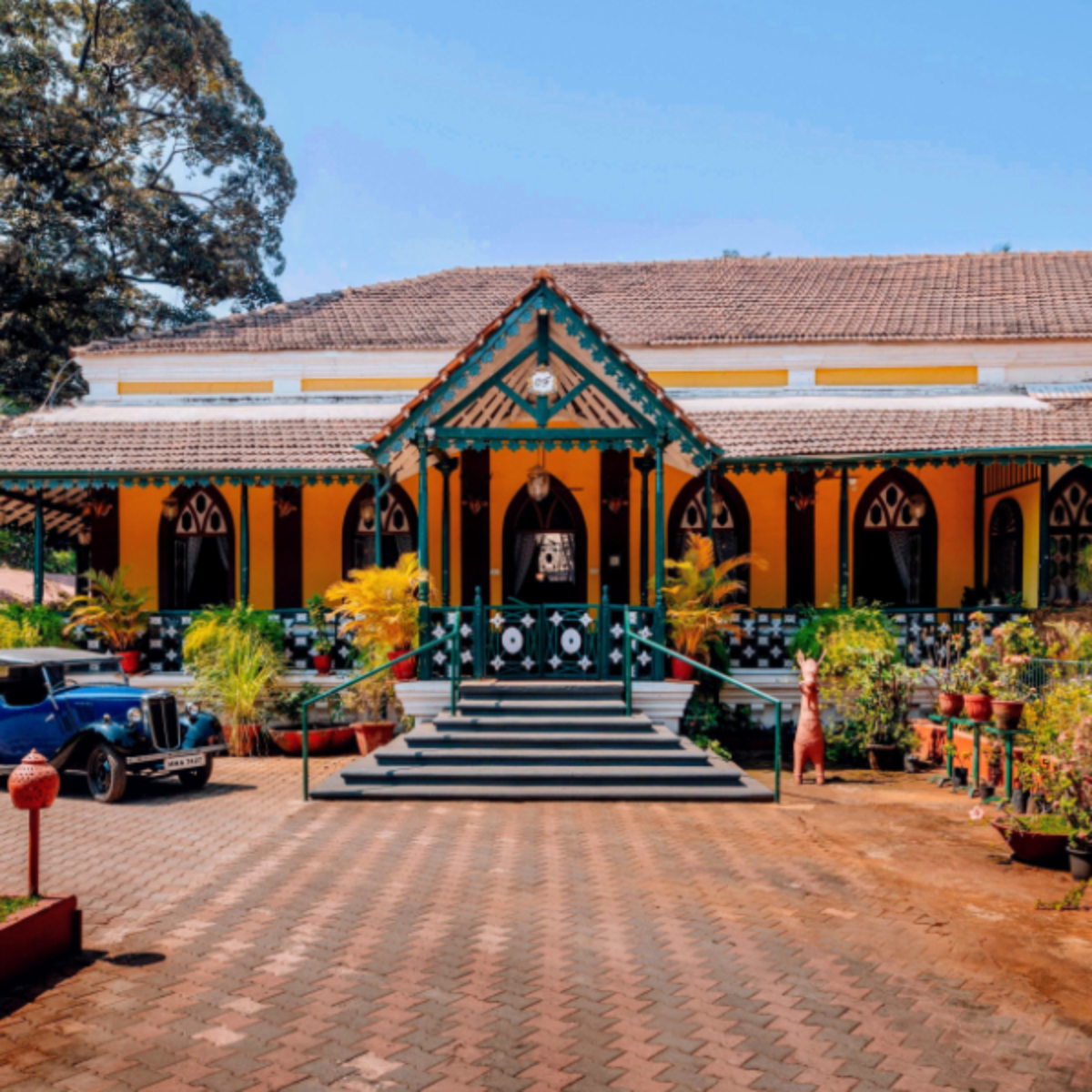 Yellow colonial-style building with teal trim and a central covered entrance, surrounded by potted plants and a vintage car to the left.