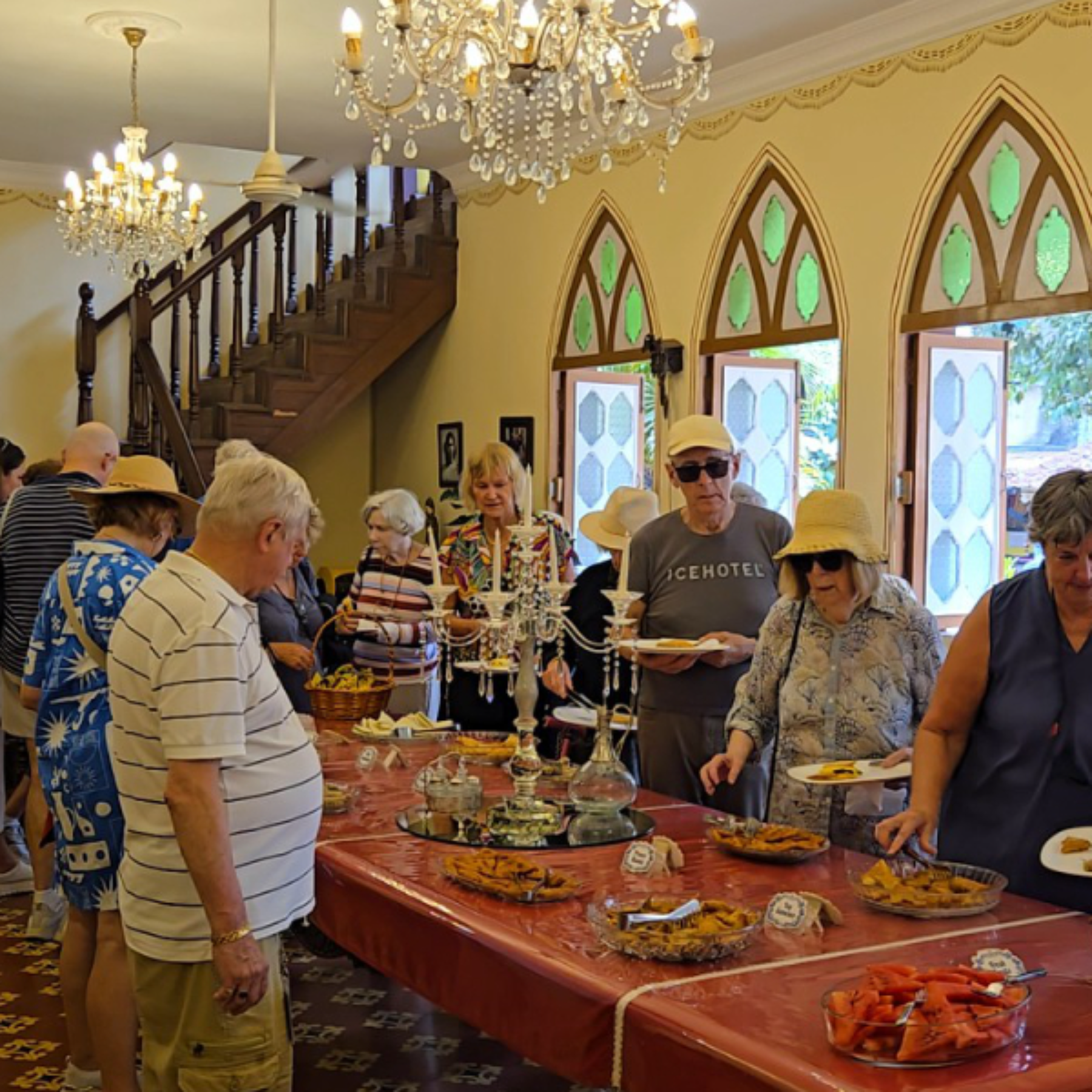 Group of seniors gathered around a long table of desserts in a warmly lit hall with chandeliers and arched windows also showing a staircase in the background to the left.