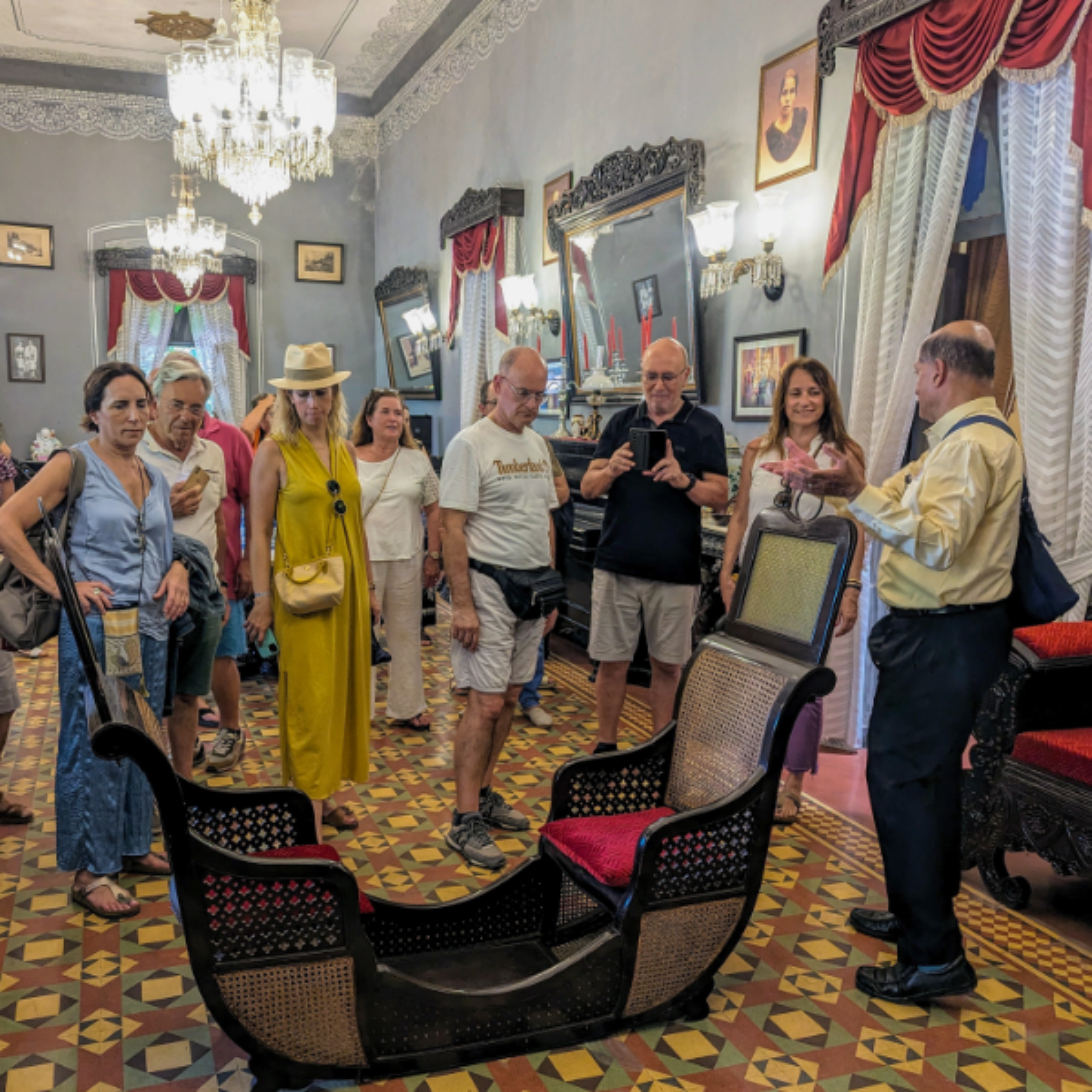 Group of tourists in a vintage showroom admiring carved chairs and ornate decor with chandeliers and patterned tile floor.