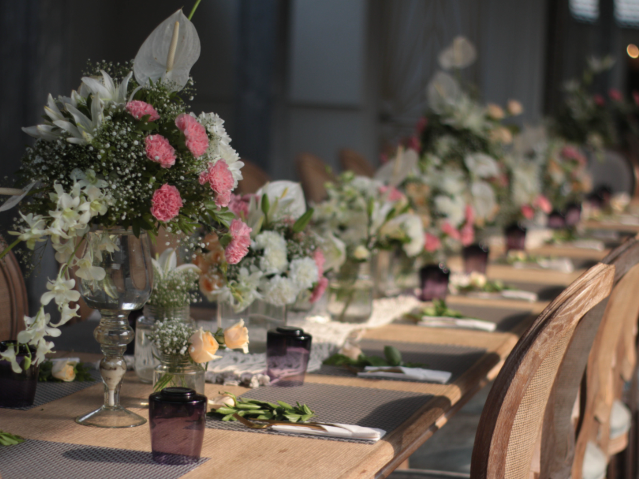 Long row of floral centerpieces in glass vases with pink and white flowers along a rustic wooden table at an event venue