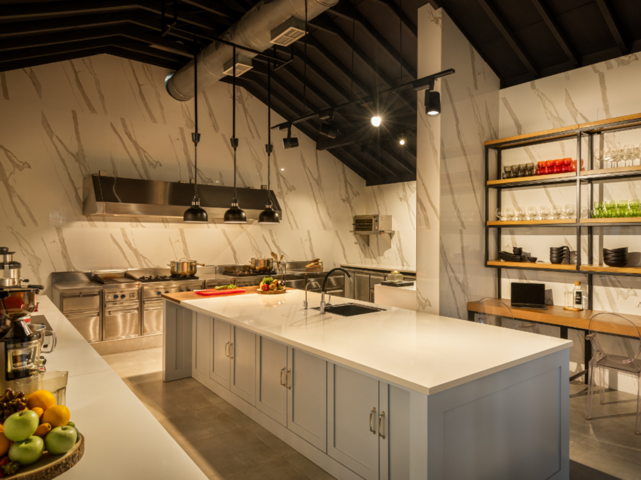 Modern open kitchen with marble walls, stainless-steel appliances, black pendant lights, and open shelving with colored glassware.