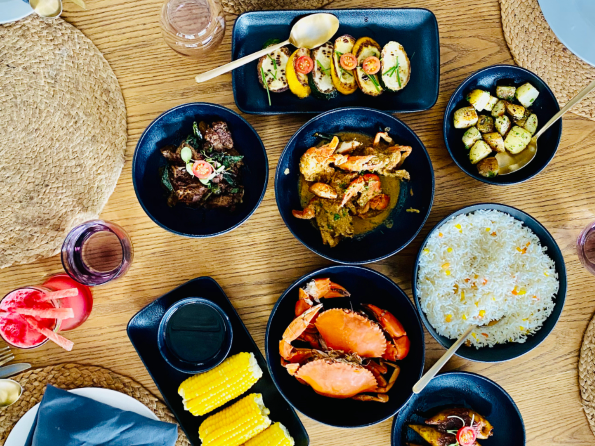 A table spread of seafood and vegetables in dark bowls: lobster, shrimp, grilled zucchini and corn alongside rice on a wooden table.