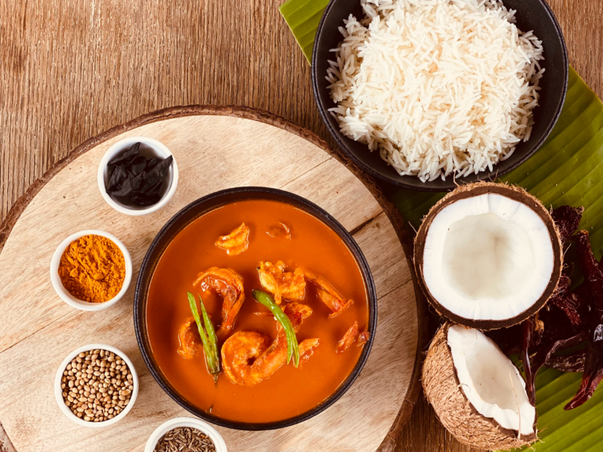 Pot of shrimp curry with green beans, served with white rice on a wooden table featuring spice bowls and coconutsCut in half beneath a rustic spread of Indian spices and rice