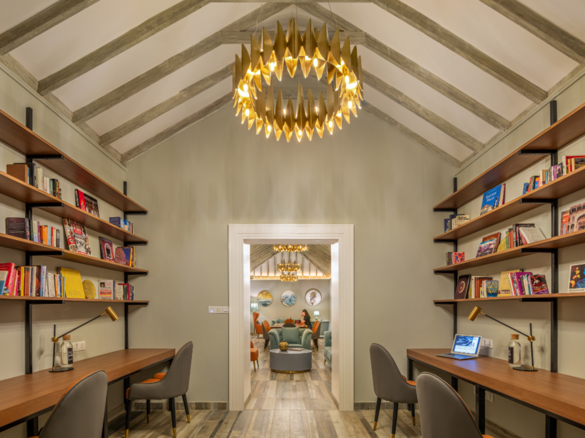 Shelf-lined study area with long wooden desks and gray chairs, a golden geometric chandelier hanging from a vaulted ceiling, and a doorway to a cozy seating area beyond.
