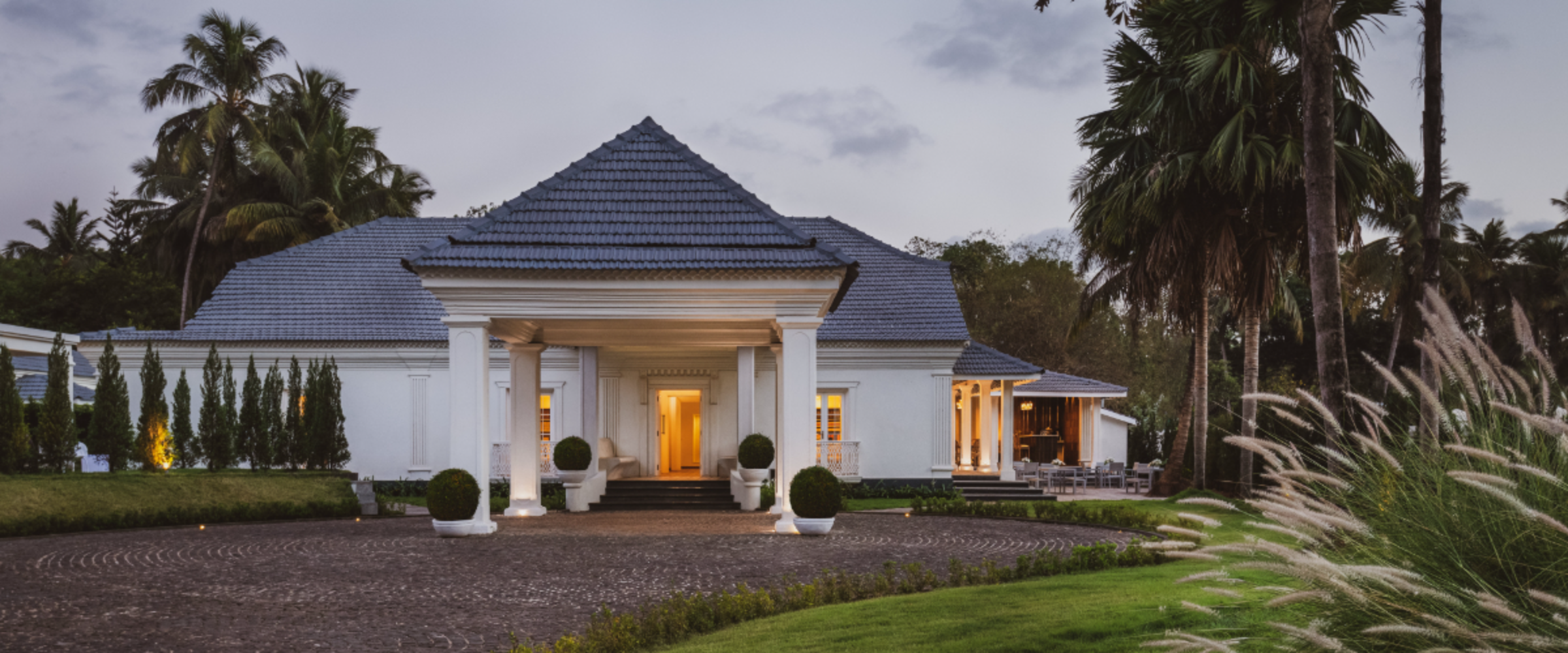 White villa with a blue-tiled roof, illuminated entrance, and palm trees at dusk.