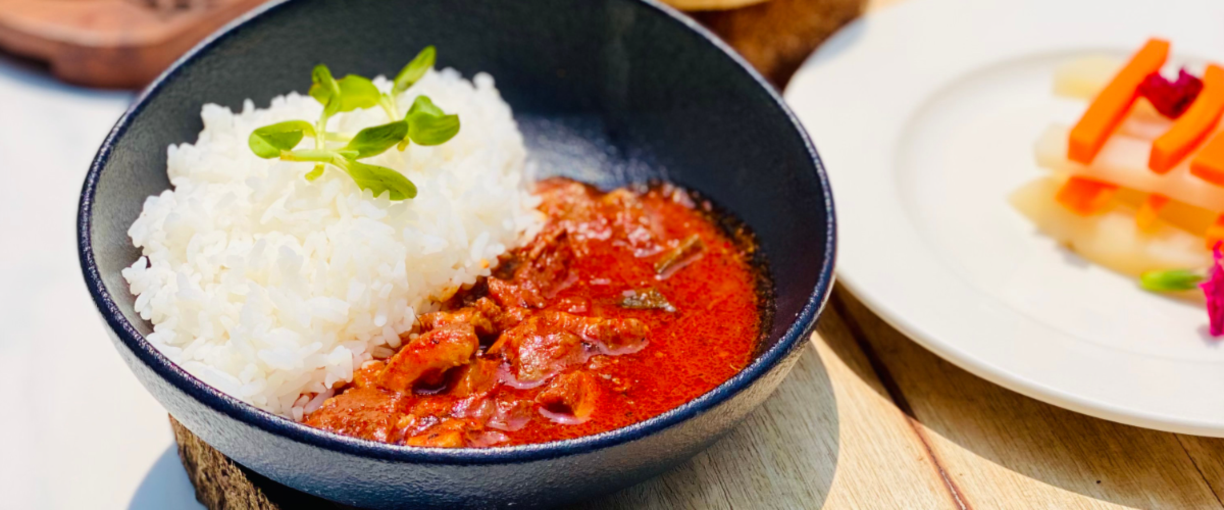 Bowl of white rice with a side of red curry in a black bowl, garnished with greens on top, placed on a wooden surface.