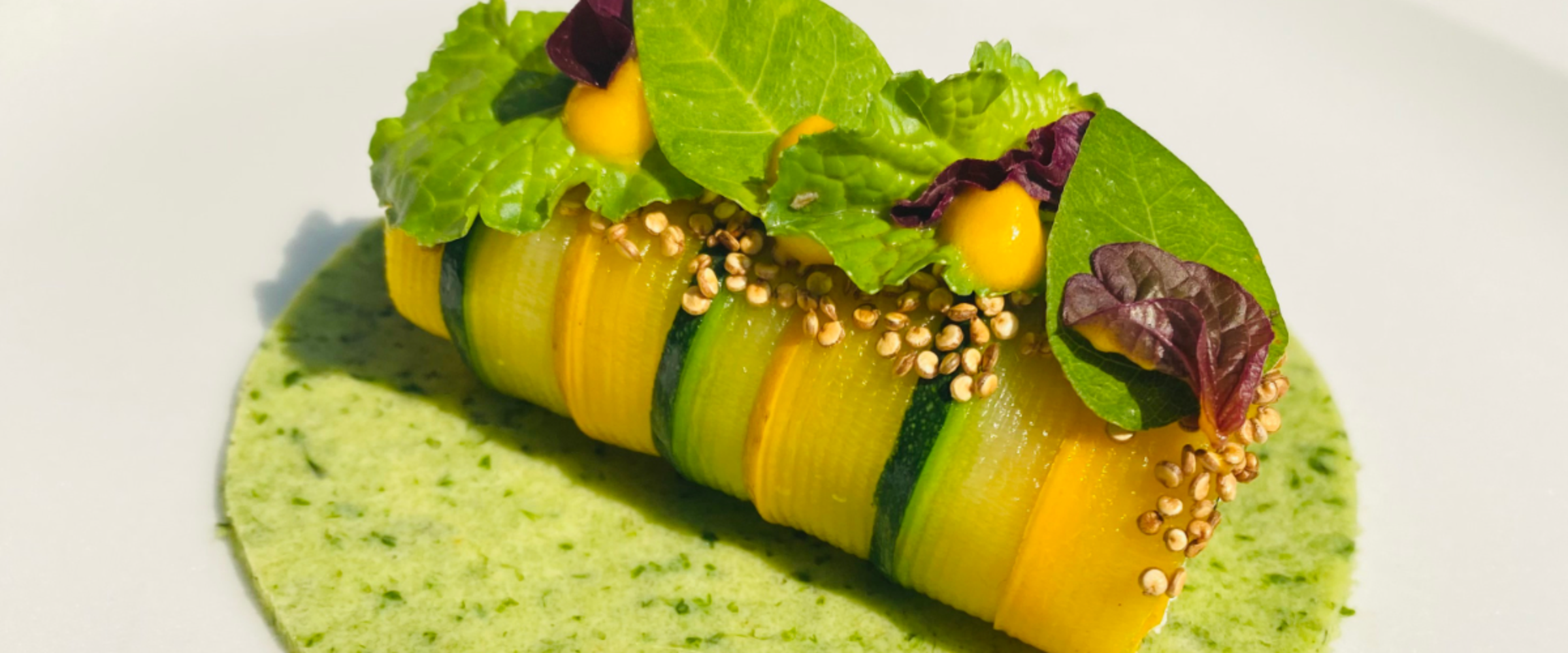 Stack of yellow and green vegetable ribbons topped with leafy greens and sesame seeds on a green herb sauce plate, a vibrant appetizer. (informative)