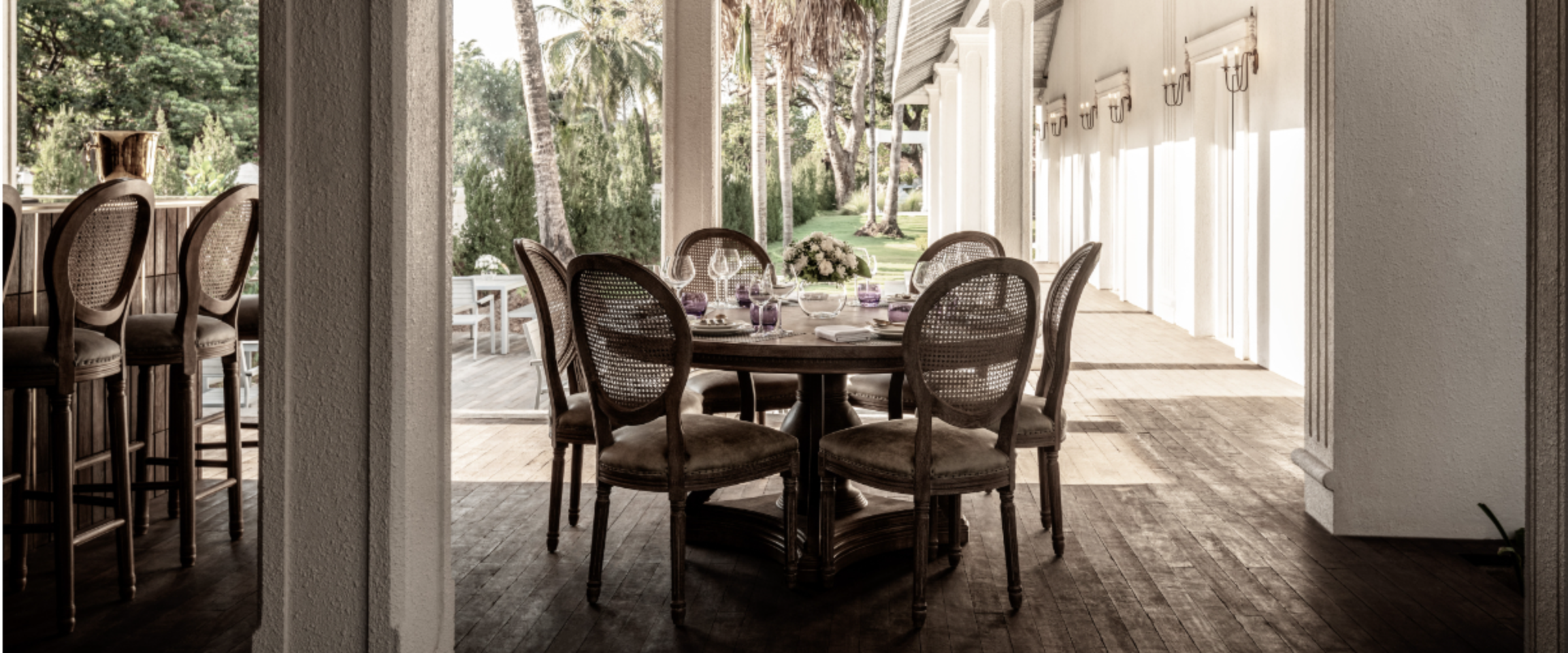 Round dining table with six wicker chairs on a covered patio, set with flowers, glassware, and purple napkins, overlooking a green garden.