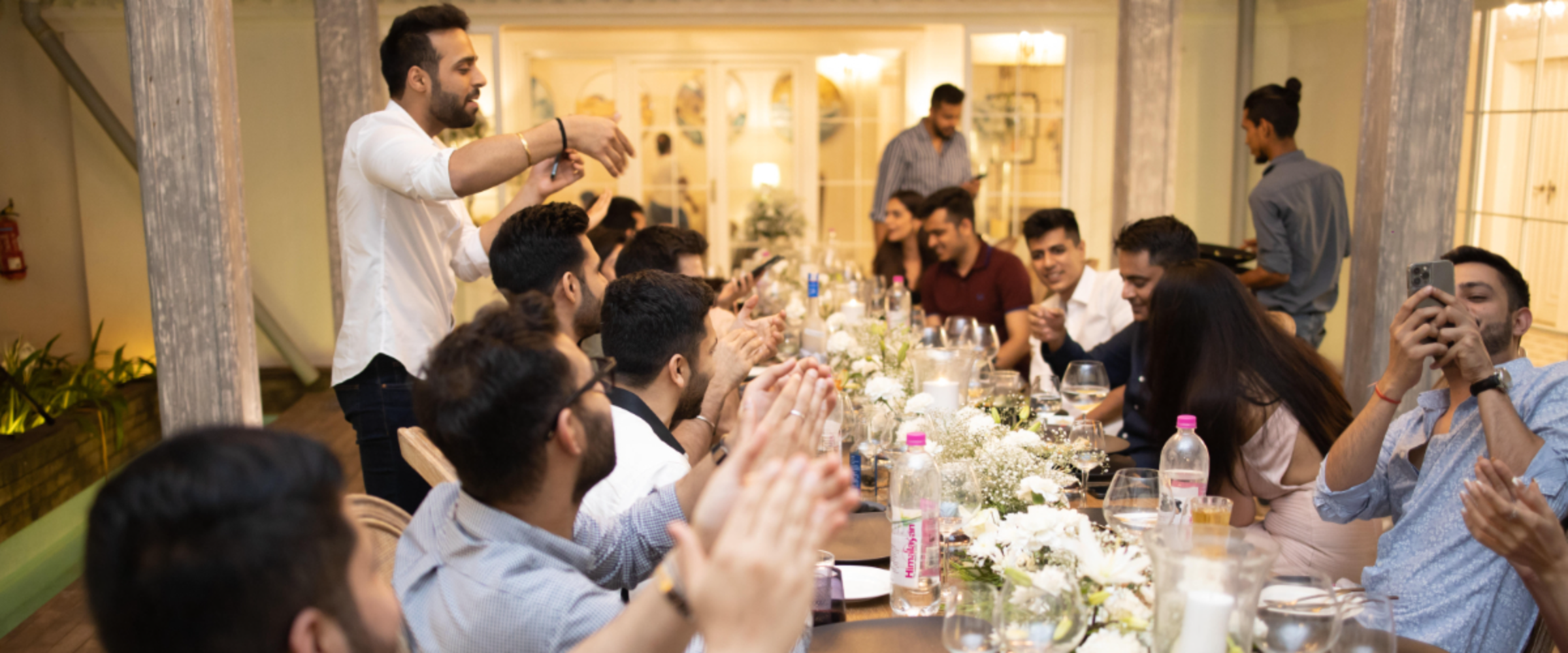 A group of friends at a long dinner table claps as a man standing at the end appears to give a toast or talk, in a warmly lit indoor setting.