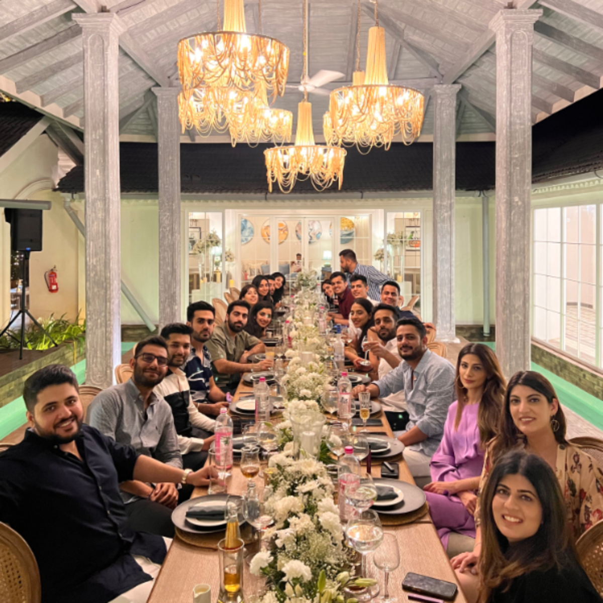 Large group of people dining together at a long banquet table under ornate chandeliers in a bright, elegant venue.