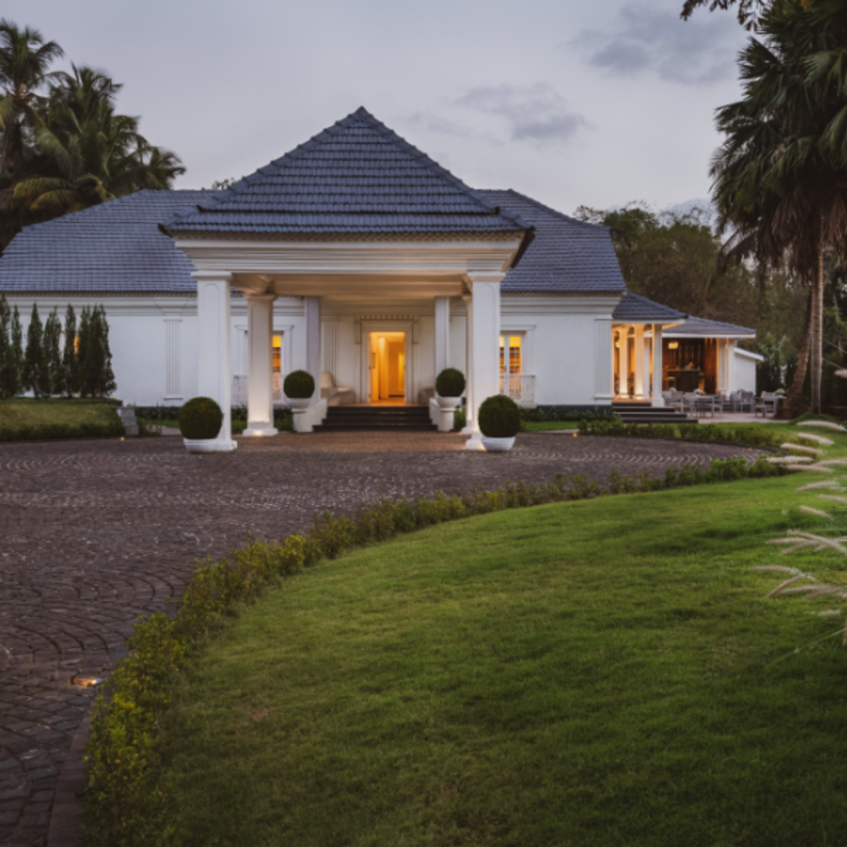 White luxury villa with a blue-tiled roof, grand portico, and warm entrance lights at dusk, surrounded by palm trees and a curved driveway.