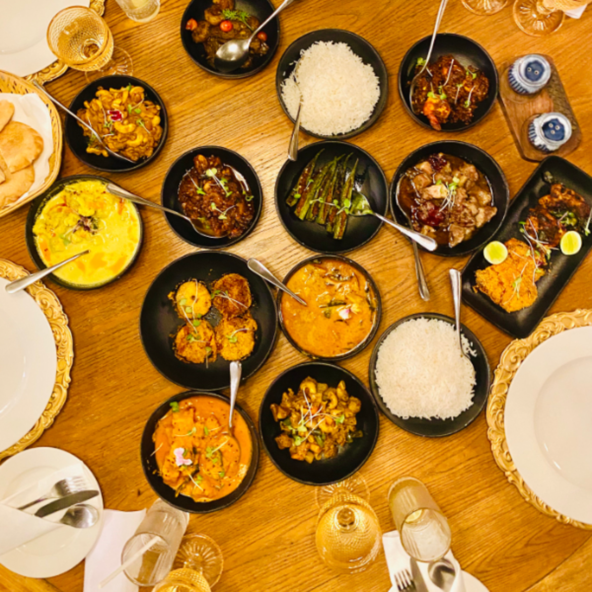 Overhead view of a colorful family-style feast with bowls of rice, curries, and vegetables around a wooden table.