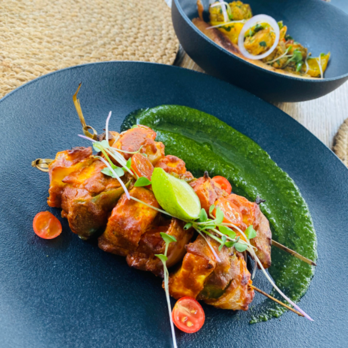 Skewered grilled chicken tikka with lime, microgreens, and a green chutney on a dark blue plate, garnished with cherry tomatoes.