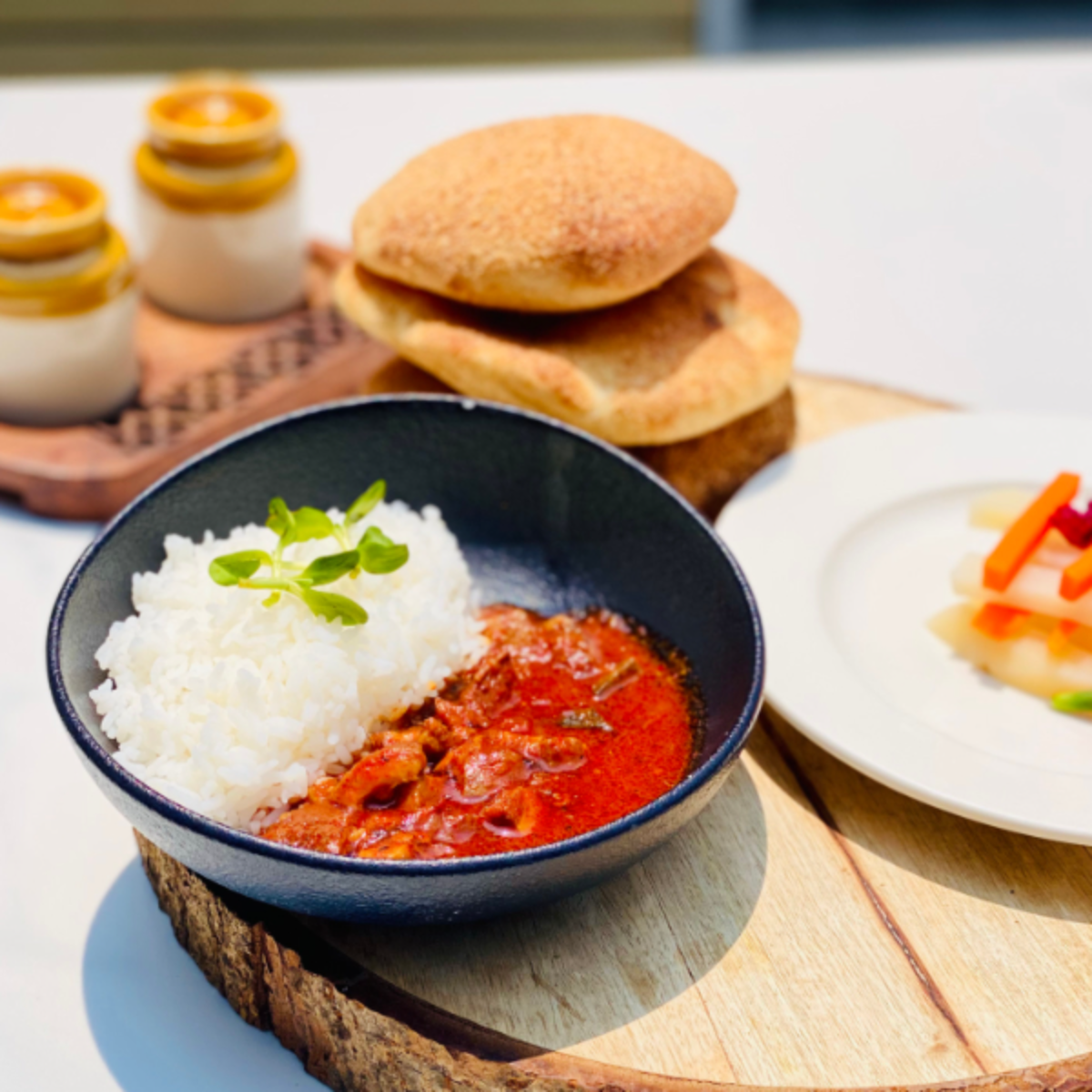 Bowl of white rice with red curry, garnished with a green herb, on a wooden board with bread rolls in the background.