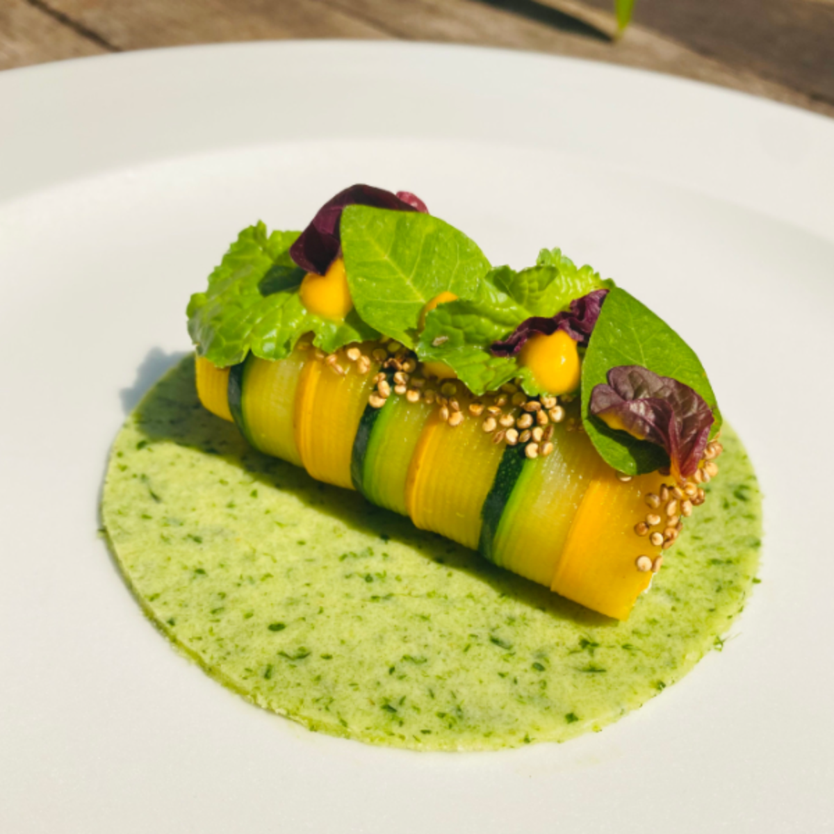 Zucchini and yellow squash ribbons on a green herb sauce, topped with leafy greens and sesame seeds, on a white plate.
