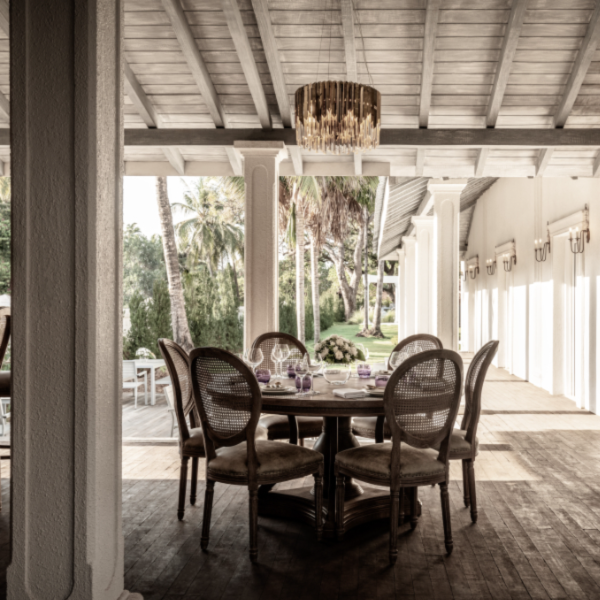Covered outdoor dining area with a round table and six wicker chairs, chandelier overhead, palm trees beyond.