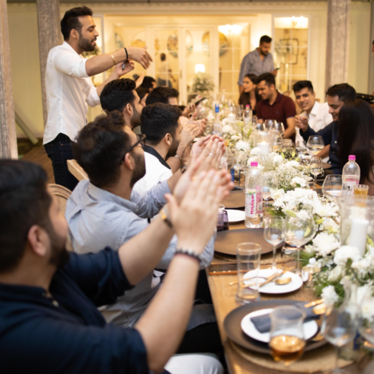 Group of friends at a long banquet table toasting and clapping during a celebration.