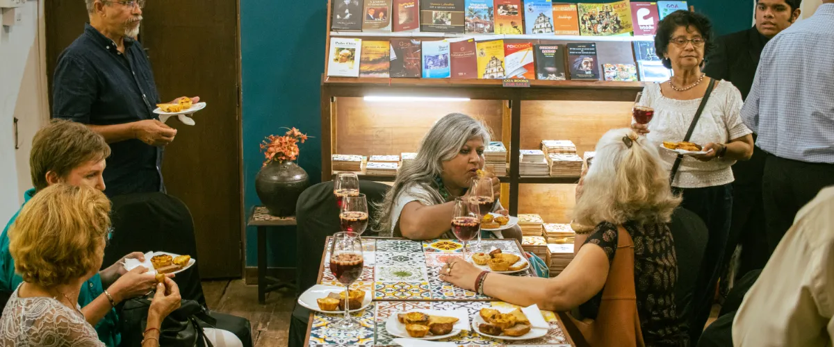 Group of adults sharing pastries and wine at a bookstore cafe, around a tiled table, with bookshelves in the background.