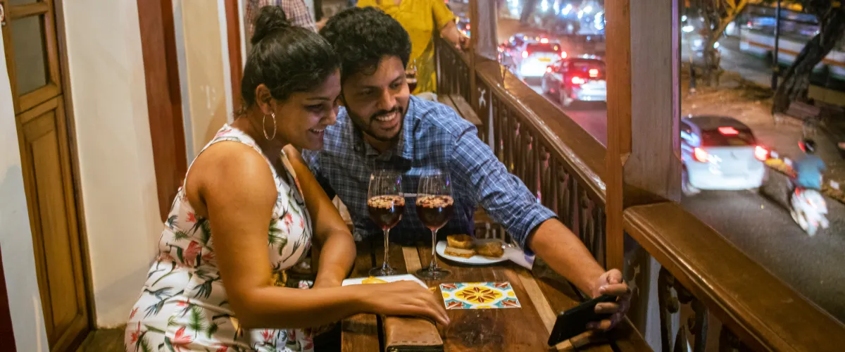 Couple sitting at a wooden balcony table, taking a selfie with a smartphone, with two glasses of red wine and dessert plates nearby on the table at night.