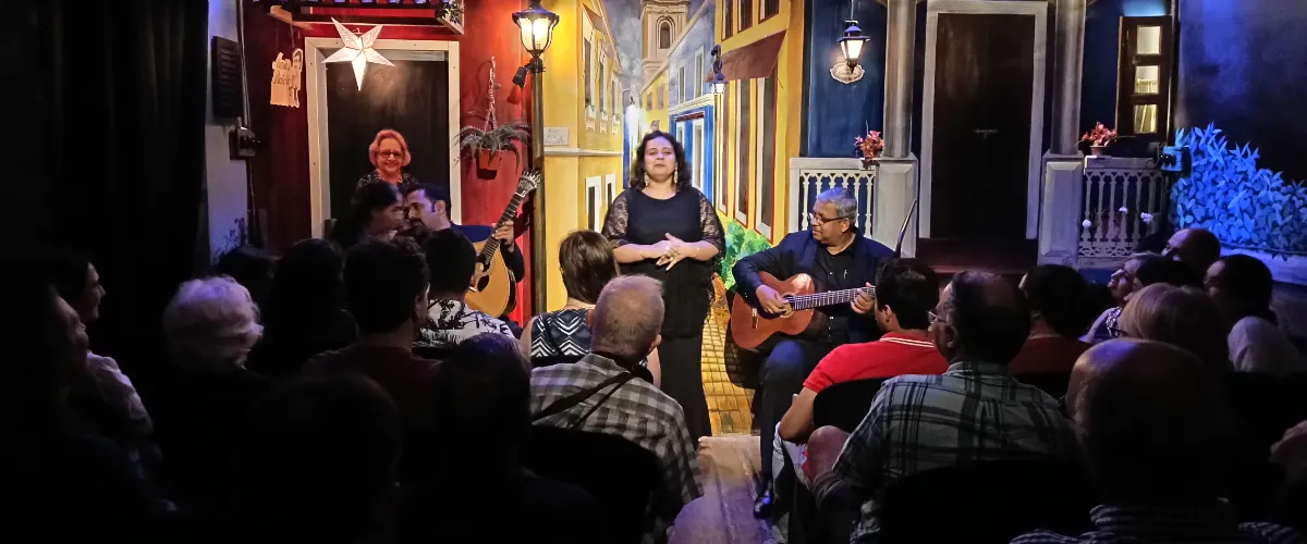 Small stage with a woman singing center stage while two guitarists accompany her for a seated audience in a colorful alleyway set.