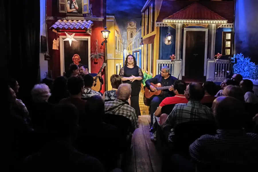 Singer center stage with two guitarists and a seated audience in a colorful painted street backdrop.